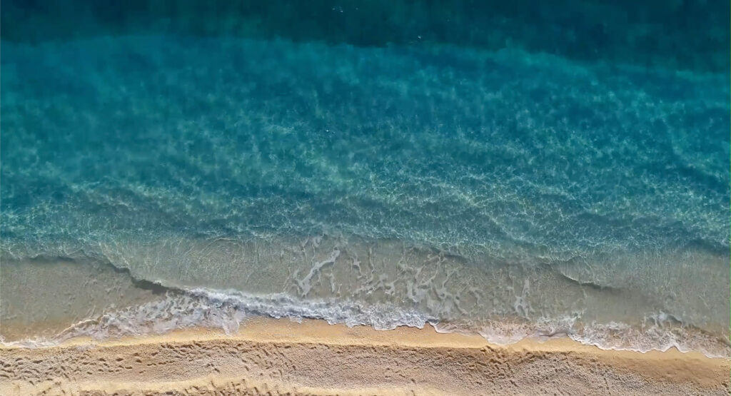 Vista aérea de una playa con aguas cristalinas y arena suave, donde las olas rompen suavemente en la orilla. Sobrevive abusos Opus Dei