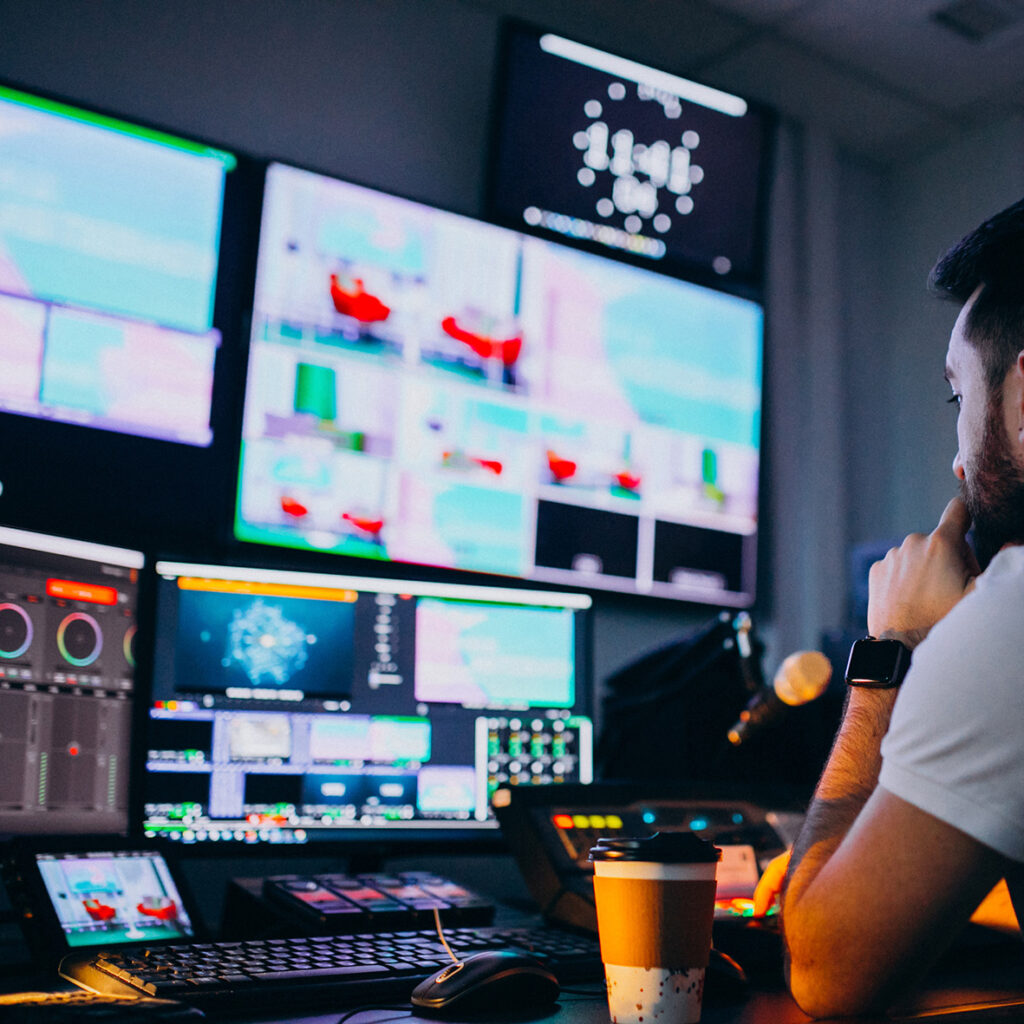 Un operador de video trabajando en un estudio, rodeado de múltiples pantallas que muestran diferentes imágenes y gráficos, mientras sostiene una taza de café. Forma que trabajamos abusos