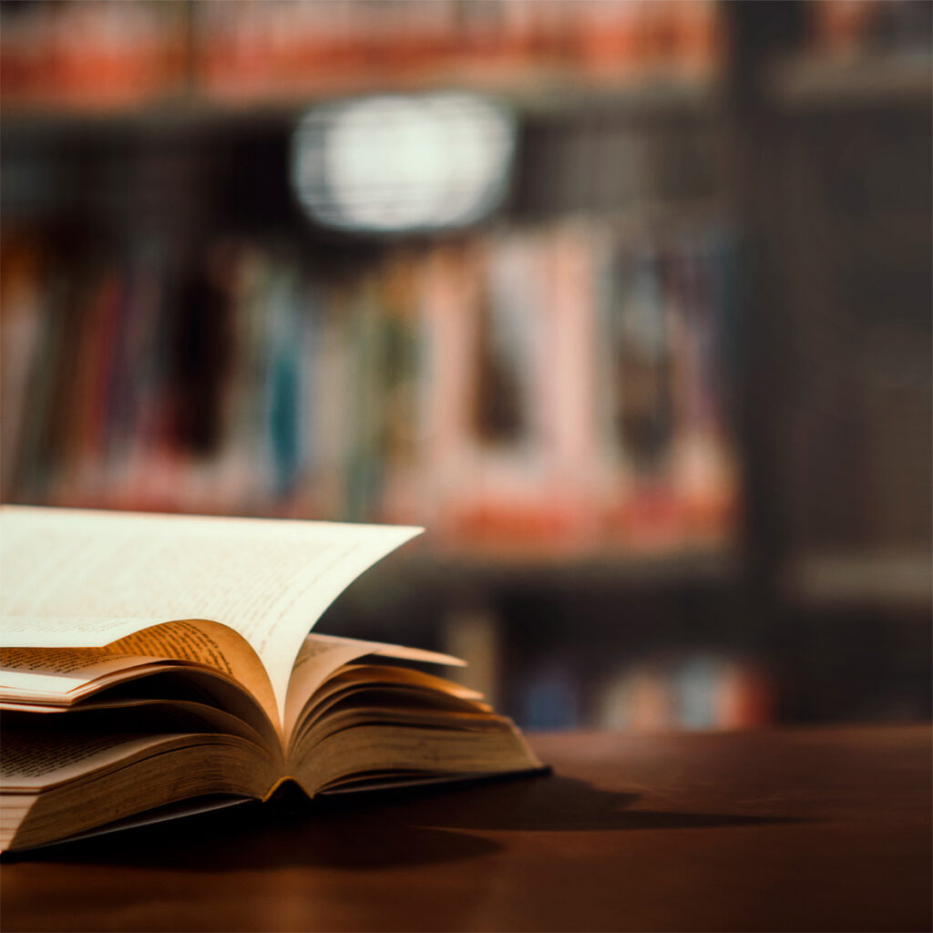 Un libro abierto sobre una mesa, con estantes llenos de libros desenfocados al fondo. Colabora 