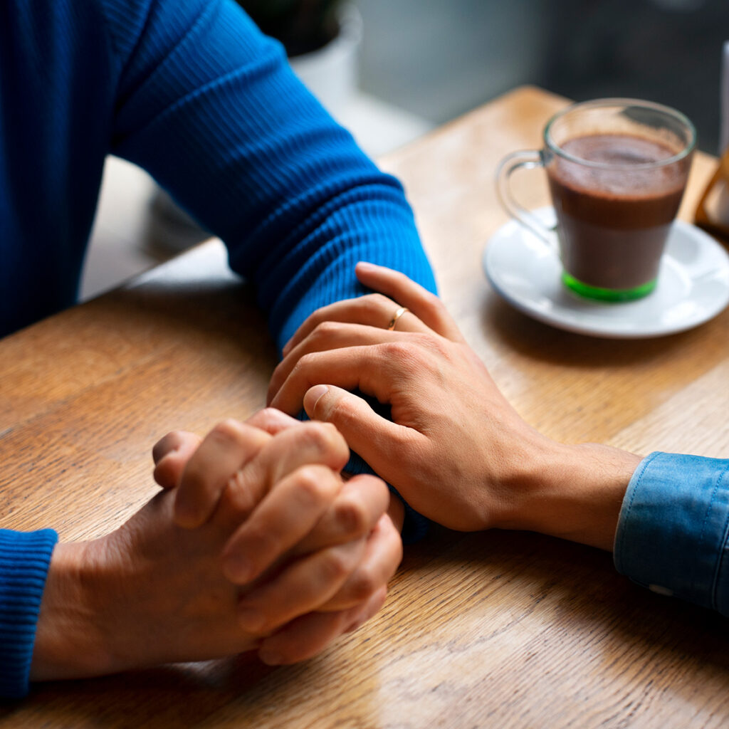 Manos entrelazadas de dos personas, una con un anillo y la otra en posición de apoyo, sobre una mesa de madera. En el fondo, una taza de café con un plato. Ayuda reconstrucción acompañamiento